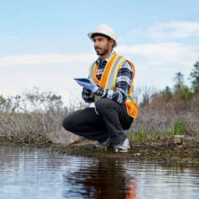 Photo of a man wearing a hard hat sitting by the water with a notepad in his hand.