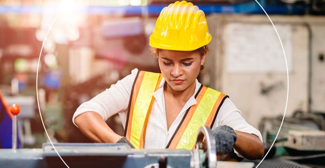 Photo of a woman wearing a work vest and hardhat working machinery 