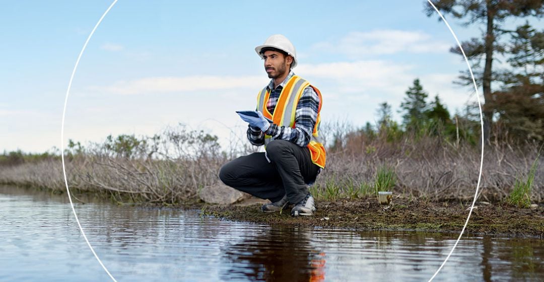 Photo of a man wearing a hard hat sitting by the water with a notepad in his hand.